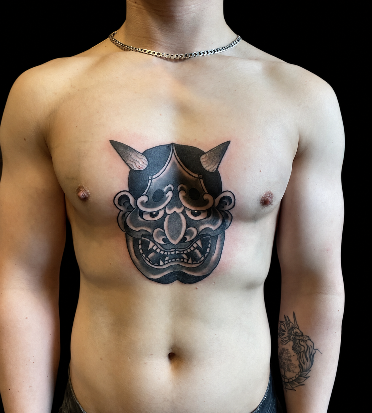 Black Japanese mask chest tattoo displayed on a black gallery background